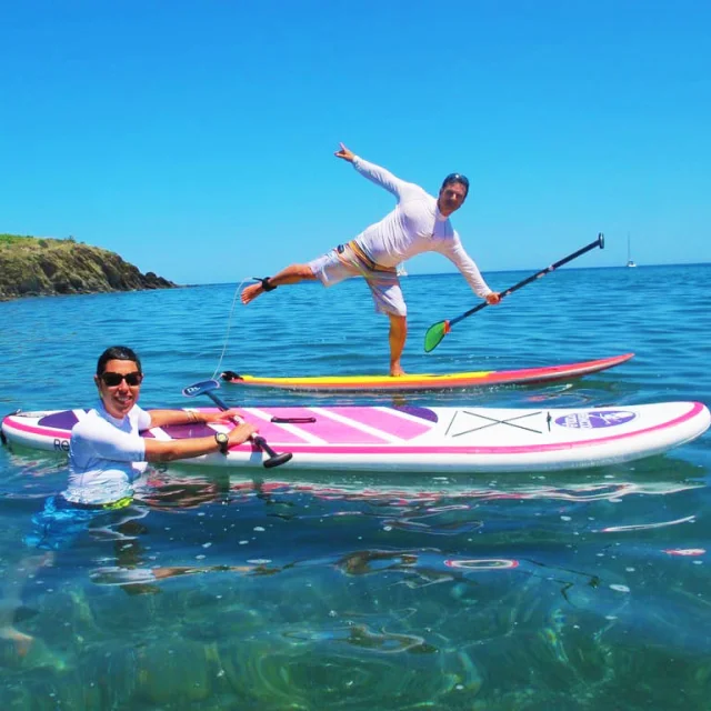 Deux personnes s’amusent sur leurs planches de stand up paddle dans une mer turquoise, sous un ciel bleu éclatant, au large des criques rocheuses de la Côte Vermeille.