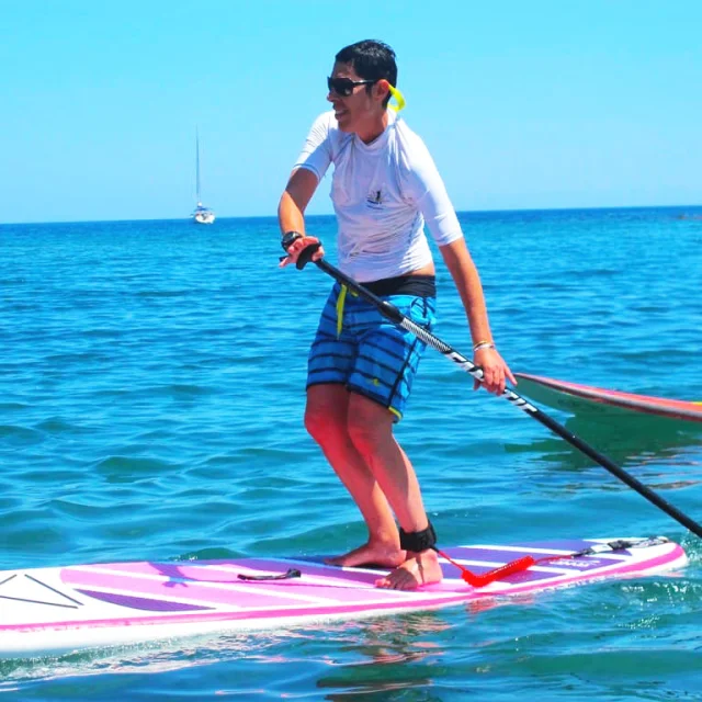 Personne pratiquant le stand up paddle sur une planche rose, pagayant sur la mer calme sous un ciel bleu, avec un voilier à l’horizon.