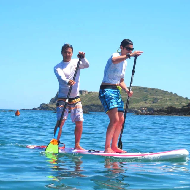 Deux personnes pratiquent le stand up paddle sur une mer calme et turquoise, au large d’un littoral verdoyant sous un ciel bleu d’été.