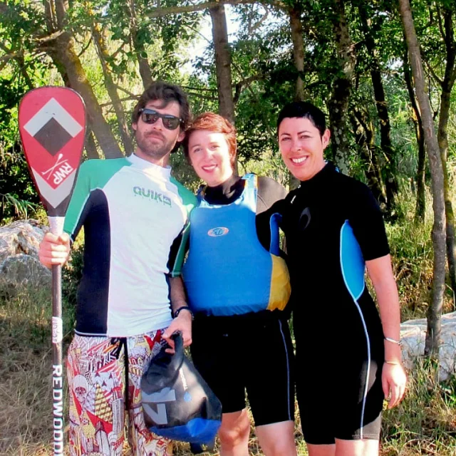 Trois personnes en tenue de stand up paddle sourient face à la caméra, équipées de gilets et de pagaies, dans un cadre naturel bordé d’arbres.