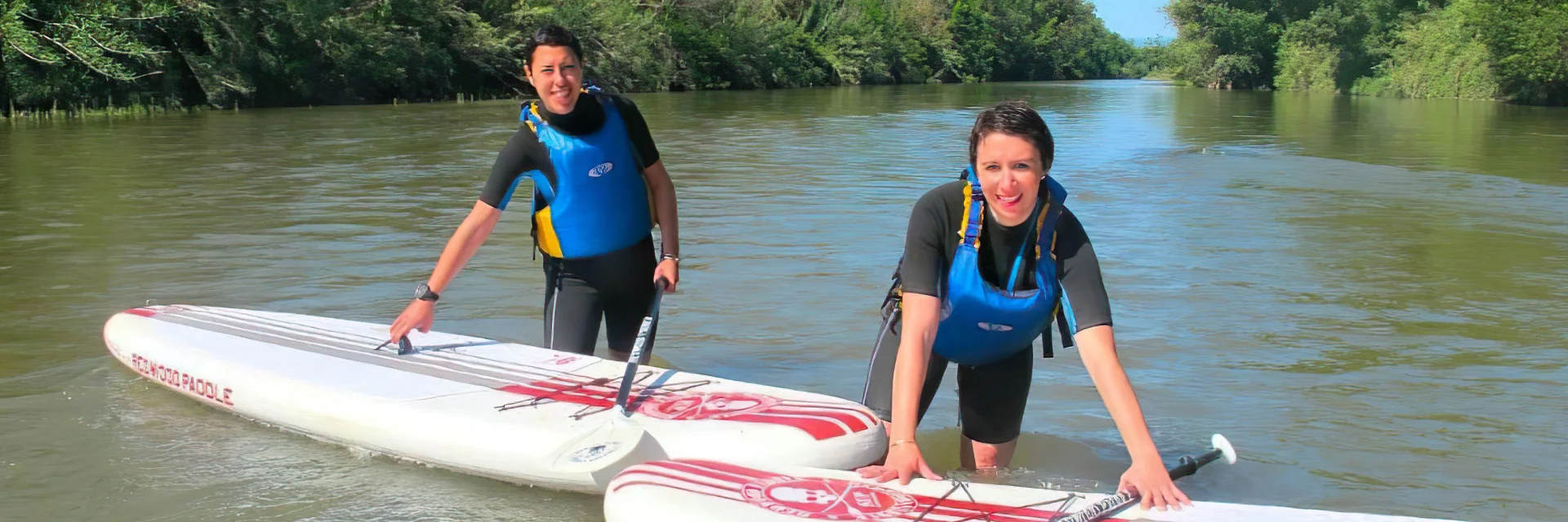 Deux personnes en combinaison et gilet de sauvetage pratiquent le stand up paddle sur une rivière bordée d’arbres.