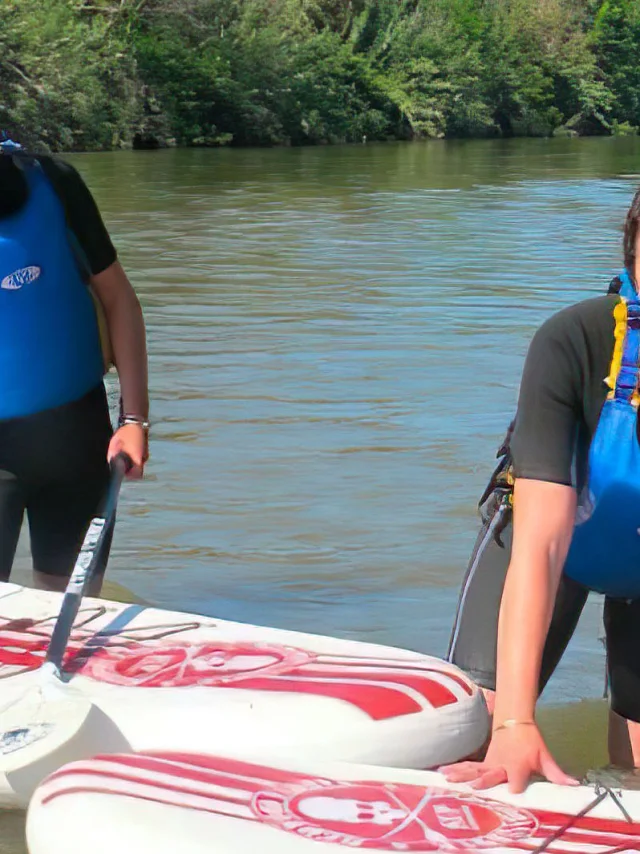 Deux personnes en combinaison et gilet de sauvetage pratiquent le stand up paddle sur une rivière bordée d’arbres.