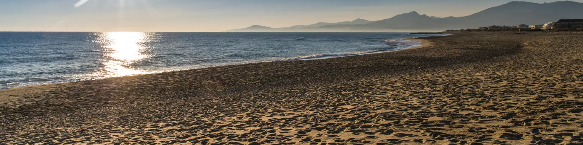 Plage de sable doré baignée par le soleil couchant, avec vue sur la mer Méditerranée et les reliefs du massif du Canigó à l’horizon.