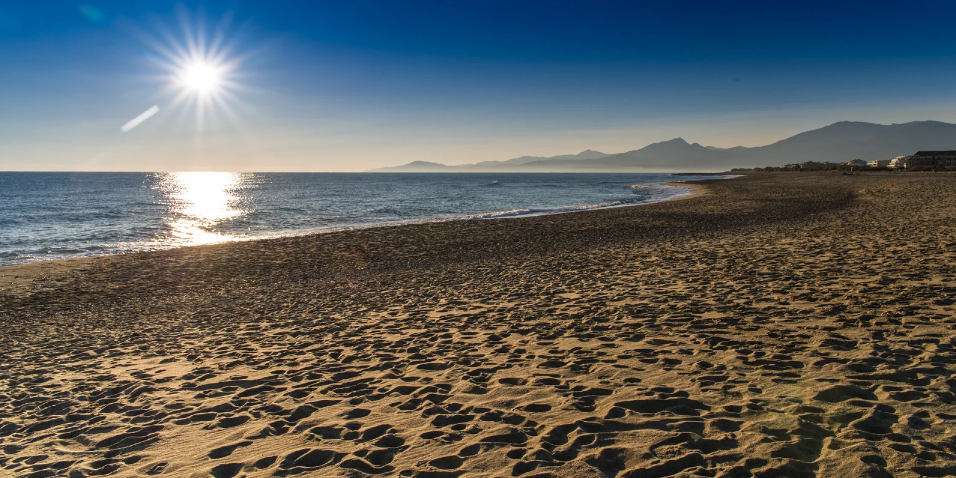 Plage de sable doré baignée par le soleil couchant, avec vue sur la mer Méditerranée et les reliefs du massif du Canigó à l’horizon.