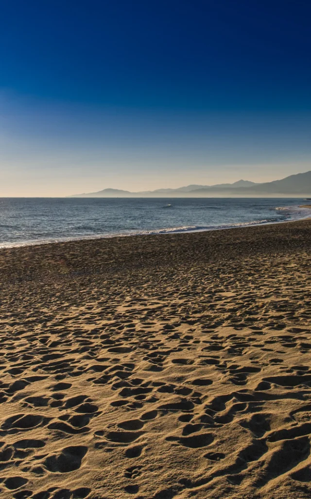 Plage de sable doré baignée par le soleil couchant, avec vue sur la mer Méditerranée et les reliefs du massif du Canigó à l’horizon.
