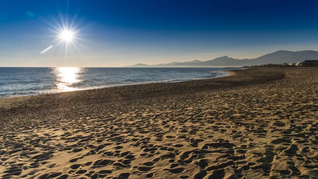 Plage de sable doré baignée par le soleil couchant, avec vue sur la mer Méditerranée et les reliefs du massif du Canigó à l’horizon.