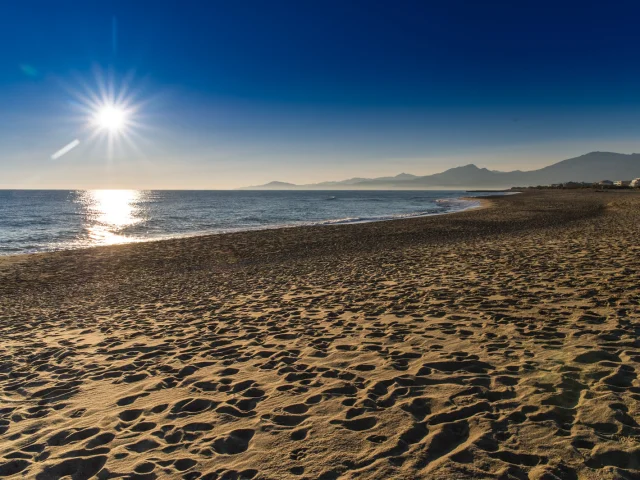 Plage de sable doré baignée par le soleil couchant, avec vue sur la mer Méditerranée et les reliefs du massif du Canigó à l’horizon.