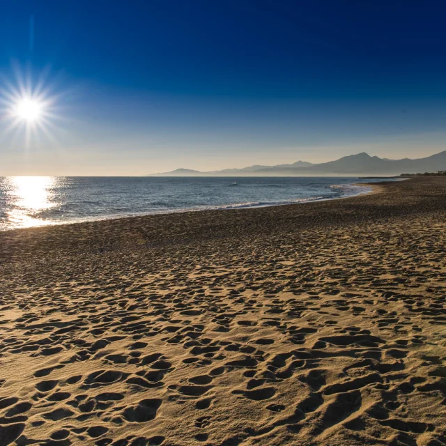 Plage de sable doré baignée par le soleil couchant, avec vue sur la mer Méditerranée et les reliefs du massif du Canigó à l’horizon.