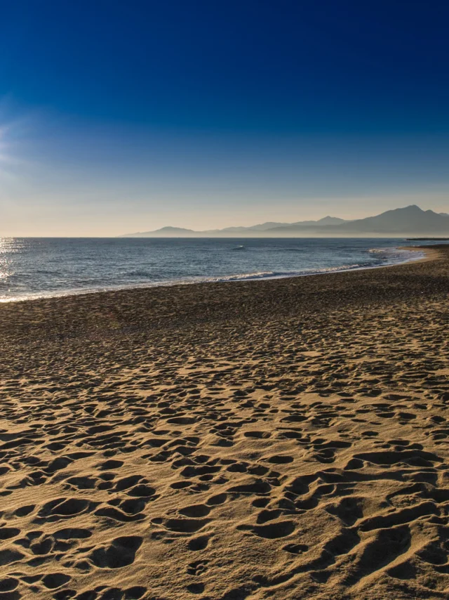 Plage de sable doré baignée par le soleil couchant, avec vue sur la mer Méditerranée et les reliefs du massif du Canigó à l’horizon.