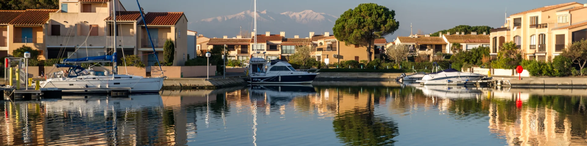 Canal bordé de maisons méditerranéennes avec des bateaux amarrés, reflet dans l’eau calme et montagnes enneigées à l’horizon.
