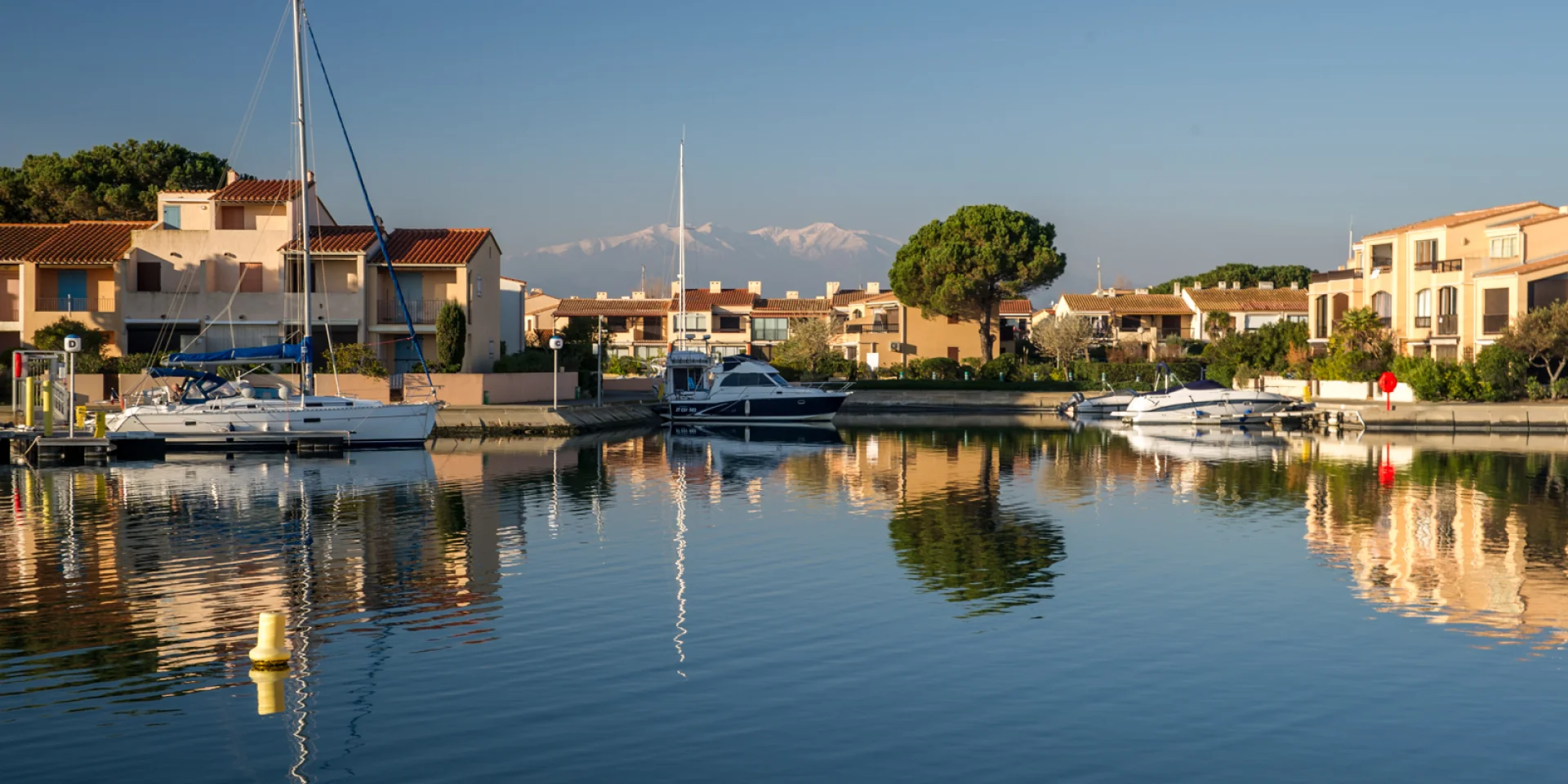 Canal bordé de maisons méditerranéennes avec des bateaux amarrés, reflet dans l’eau calme et montagnes enneigées à l’horizon.