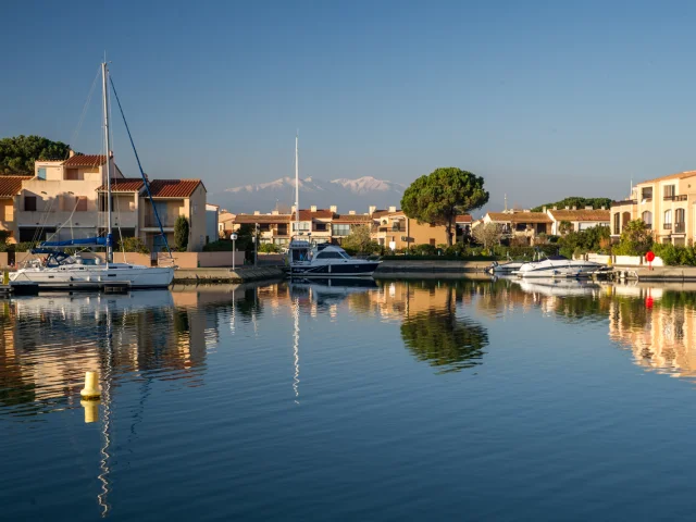 Canal bordé de maisons méditerranéennes avec des bateaux amarrés, reflet dans l’eau calme et montagnes enneigées à l’horizon.