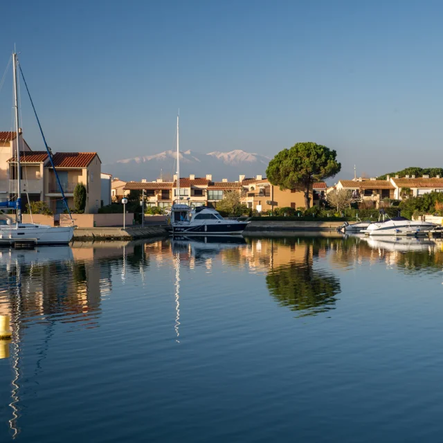 Canal bordé de maisons méditerranéennes avec des bateaux amarrés, reflet dans l’eau calme et montagnes enneigées à l’horizon.