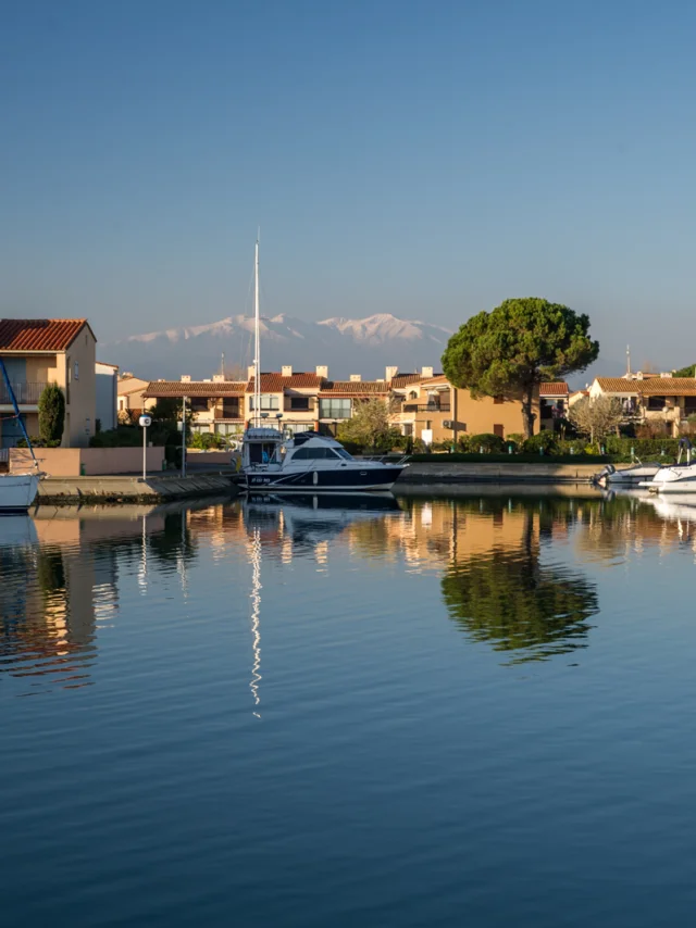 Canal bordé de maisons méditerranéennes avec des bateaux amarrés, reflet dans l’eau calme et montagnes enneigées à l’horizon.