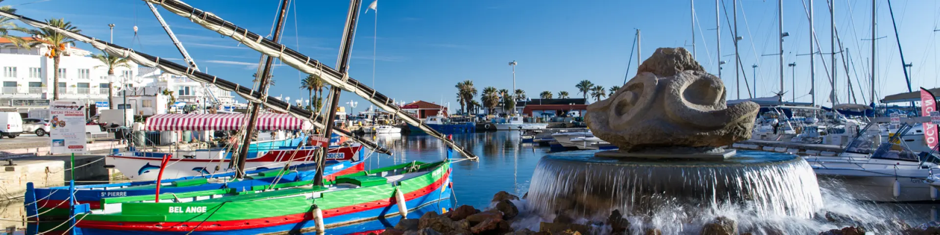 Port méditerranéen animé avec des barques catalanes colorées amarrées près d’une fontaine en pierre et des voiliers alignés sous un ciel bleu.