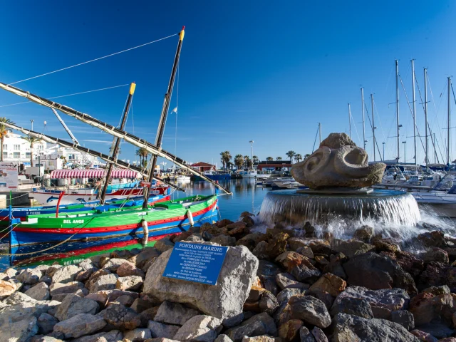 Port méditerranéen animé avec des barques catalanes colorées amarrées près d’une fontaine en pierre et des voiliers alignés sous un ciel bleu.