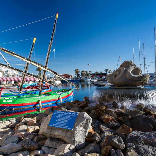Port méditerranéen animé avec des barques catalanes colorées amarrées près d’une fontaine en pierre et des voiliers alignés sous un ciel bleu.