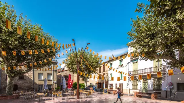 Place de village animée décorée de guirlandes aux couleurs catalanes rouge et jaune, avec terrasses de cafés ombragées sous les platanes et façades colorées.