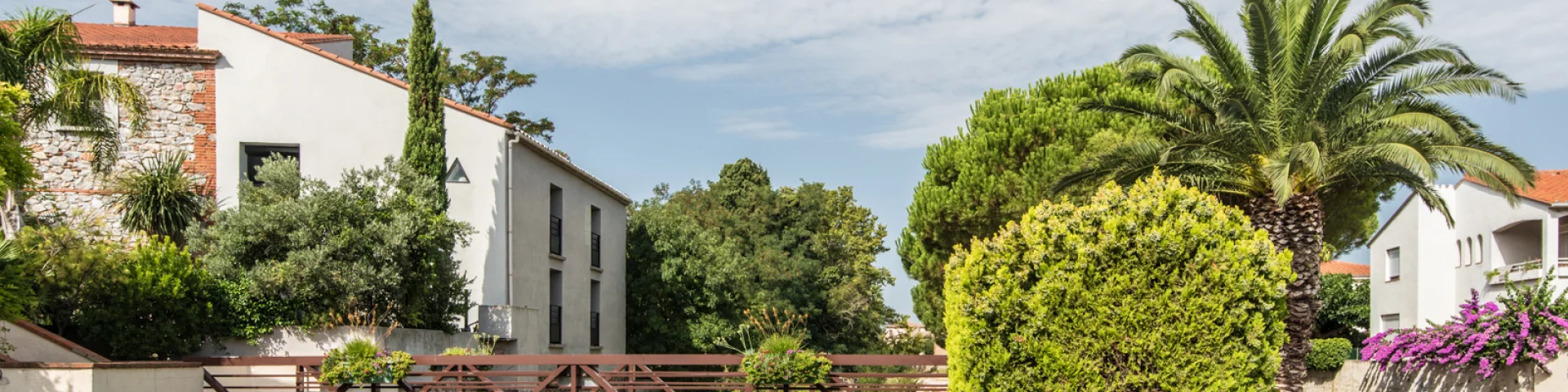 Petit pont en bois traversant un canal bordé de végétation, entouré de maisons aux façades claires et de jardins méditerranéens avec palmiers et bougainvilliers.