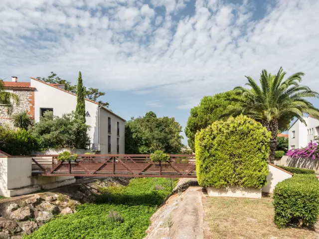 Petit pont en bois traversant un canal bordé de végétation, entouré de maisons aux façades claires et de jardins méditerranéens avec palmiers et bougainvilliers.