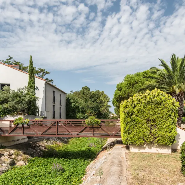 Petit pont en bois traversant un canal bordé de végétation, entouré de maisons aux façades claires et de jardins méditerranéens avec palmiers et bougainvilliers.