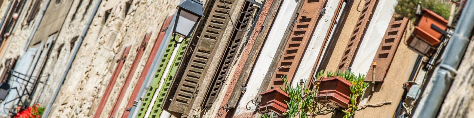 Détail des façades anciennes de Villefranche-de-Conflent, avec volets colorés, jardinières fleuries et lampadaires en fer forgé, sous le soleil catalan.