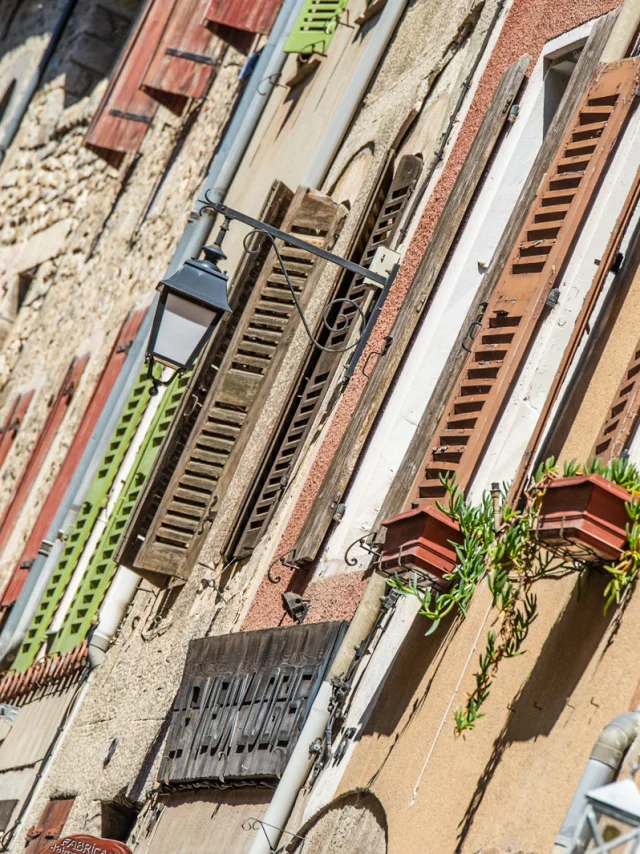 Détail des façades anciennes de Villefranche-de-Conflent, avec volets colorés, jardinières fleuries et lampadaires en fer forgé, sous le soleil catalan.