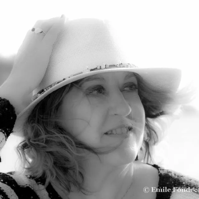 Portrait en noir et blanc d’une femme souriante portant un chapeau clair, regardant vers la droite, dans une lumière douce.