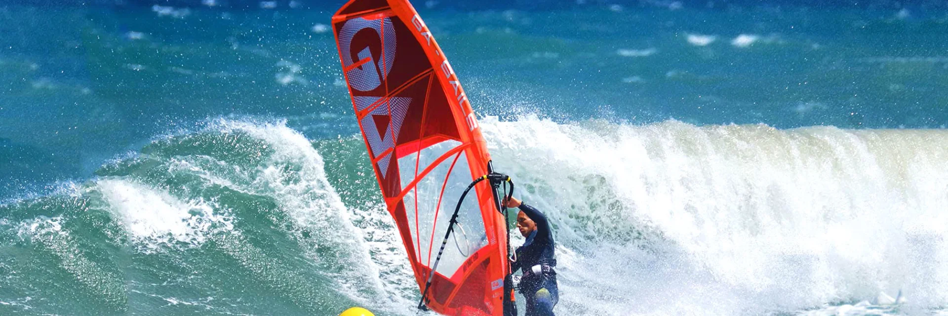 Planche à voile rouge affrontant une grande vague sur la mer Méditerranée, sous un ciel bleu éclatant.