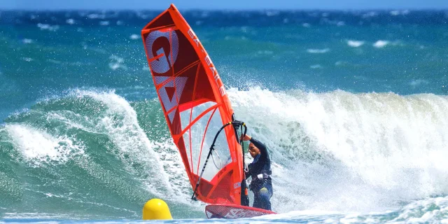 Planche à voile rouge affrontant une grande vague sur la mer Méditerranée, sous un ciel bleu éclatant.