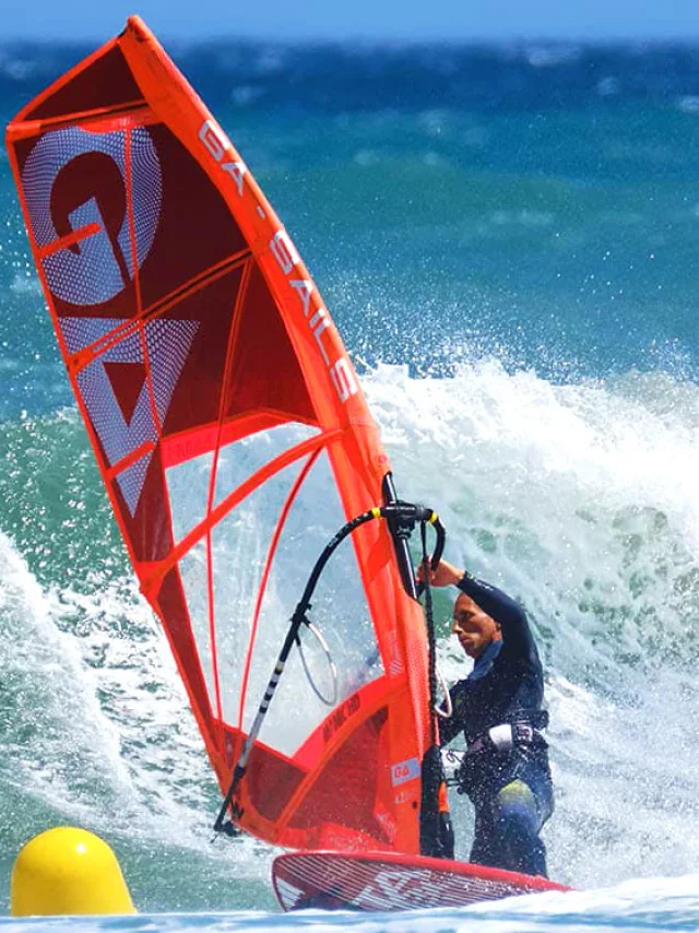 Planche à voile rouge affrontant une grande vague sur la mer Méditerranée, sous un ciel bleu éclatant.