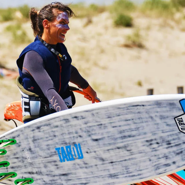 Personne souriante portant une planche de windsurf sur une plage ensoleillée, prête à rejoindre la mer.