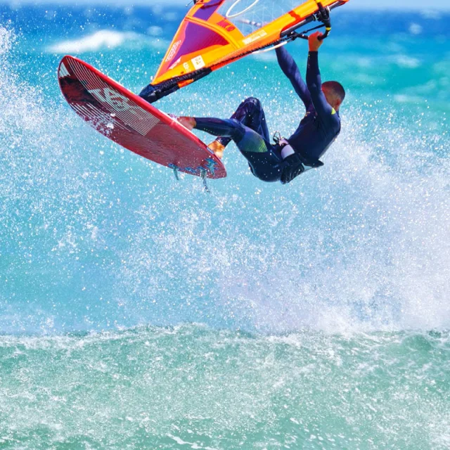 Planche à voile effectuant un saut au-dessus des vagues, voile orange et rouge, sur une mer turquoise agitée.