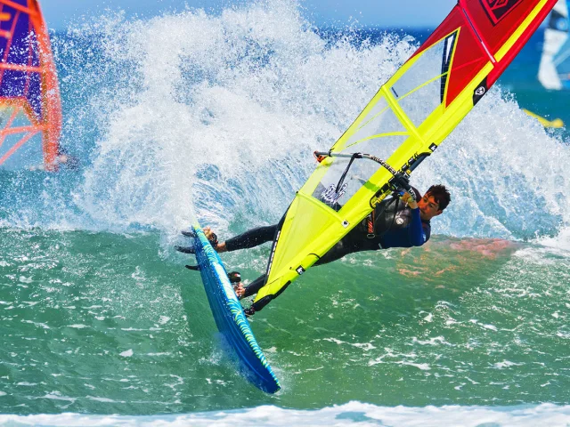 Planche à voile effectuant une figure sur une vague, voile jaune et rouge sous un ciel bleu, sur la Méditerranée.