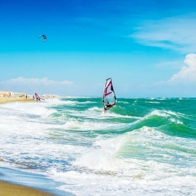 Planche à voile et kitesurf sur une plage méditerranéenne balayée par le vent, sous un ciel bleu lumineux.