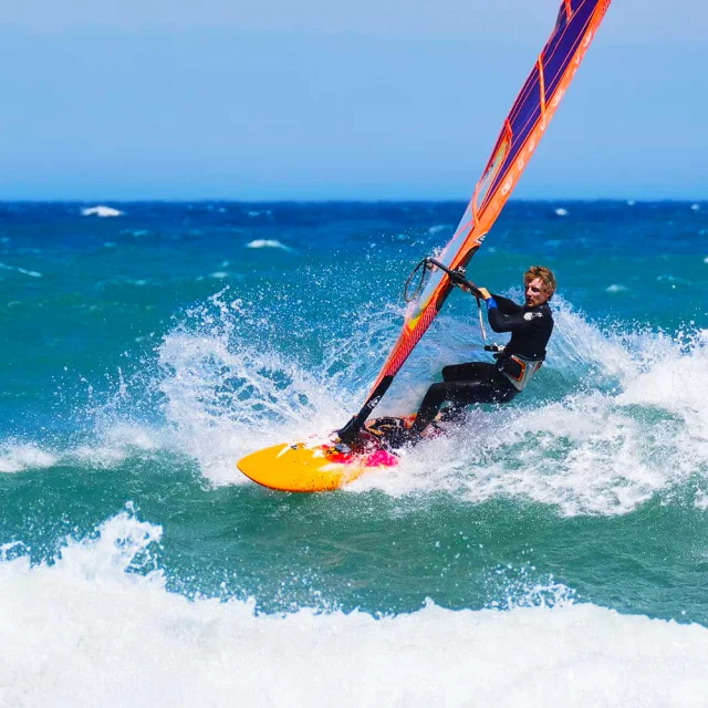 Planche à voile en action sur une vague, sous un ciel bleu et une mer turquoise agitée par le vent.