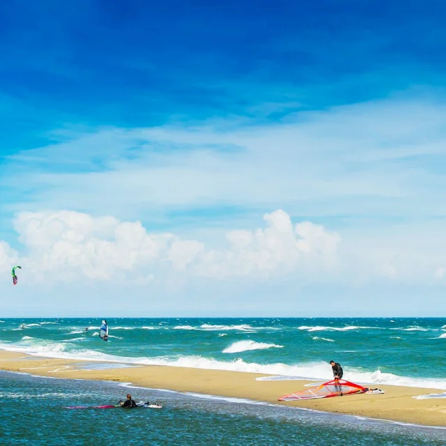 Windsurfers et kitesurfers évoluant sur une plage de sable bordée par la Méditerranée sous un grand ciel bleu.