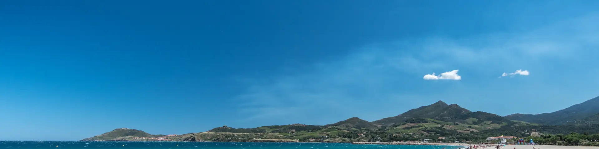 Plage de sable fin bordée par la mer turquoise à Banyuls-sur-Mer, avec des baigneurs profitant du soleil et des montagnes verdoyantes en arrière-plan.