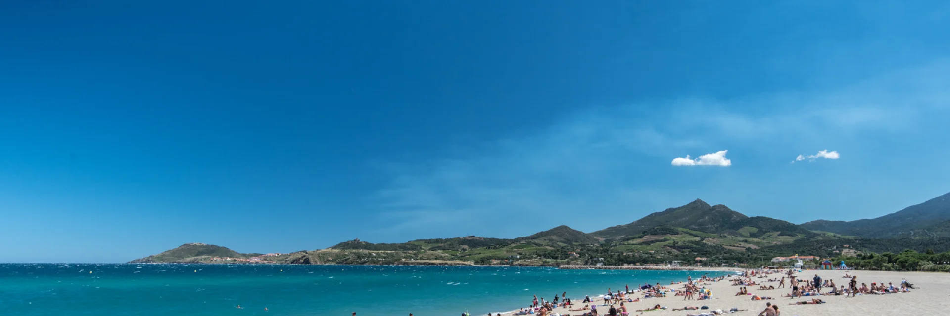 Plage de sable fin bordée par la mer turquoise à Banyuls-sur-Mer, avec des baigneurs profitant du soleil et des montagnes verdoyantes en arrière-plan.