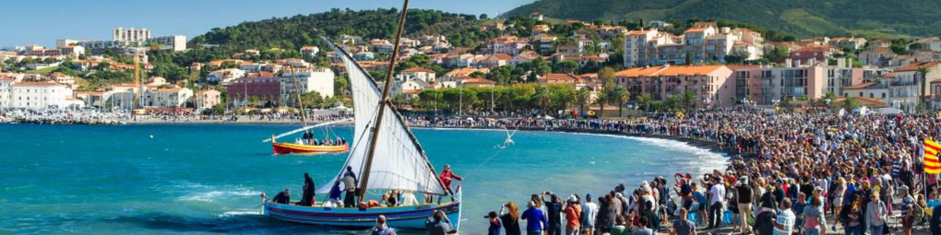 Grande foule rassemblée sur la plage de Banyuls-sur-Mer pour une fête traditionnelle avec barques catalanes à voile sur la mer.