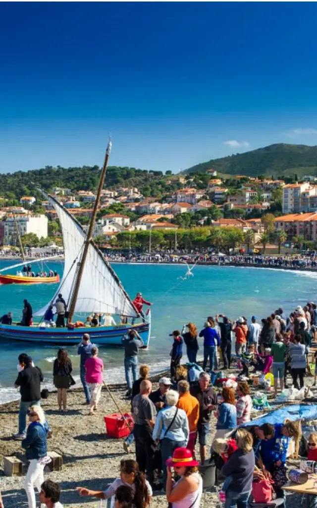 Grande foule rassemblée sur la plage de Banyuls-sur-Mer pour une fête traditionnelle avec barques catalanes à voile sur la mer.
