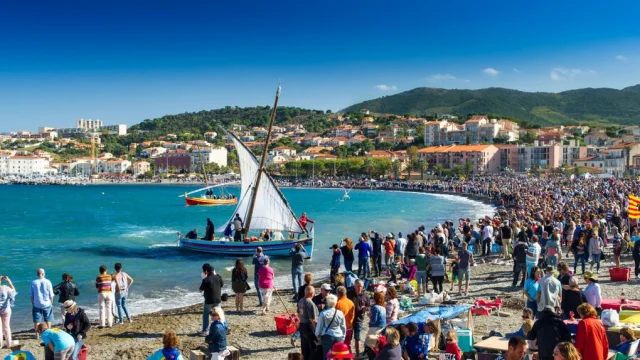Grande foule rassemblée sur la plage de Banyuls-sur-Mer pour une fête traditionnelle avec barques catalanes à voile sur la mer.