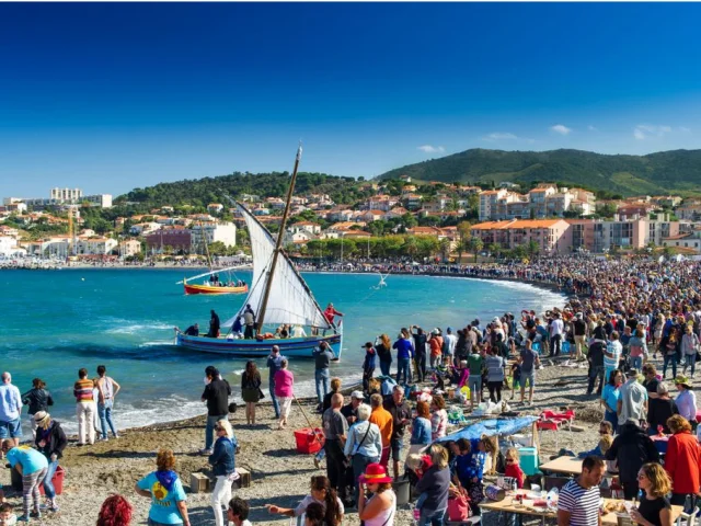 Grande foule rassemblée sur la plage de Banyuls-sur-Mer pour une fête traditionnelle avec barques catalanes à voile sur la mer.