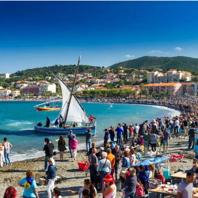 Grande foule rassemblée sur la plage de Banyuls-sur-Mer pour une fête traditionnelle avec barques catalanes à voile sur la mer.