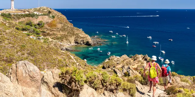 Deux randonneurs descendent un sentier escarpé dominant la mer Méditerranée, face au phare du Cap Béar et aux eaux turquoise bordées de bateaux au mouillage.