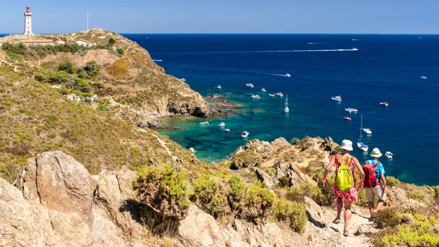 Deux randonneurs descendent un sentier escarpé dominant la mer Méditerranée, face au phare du Cap Béar et aux eaux turquoise bordées de bateaux au mouillage.