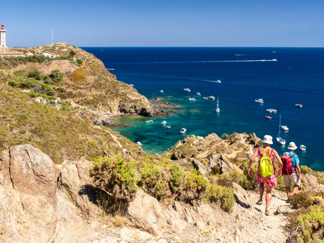 Deux randonneurs descendent un sentier escarpé dominant la mer Méditerranée, face au phare du Cap Béar et aux eaux turquoise bordées de bateaux au mouillage.