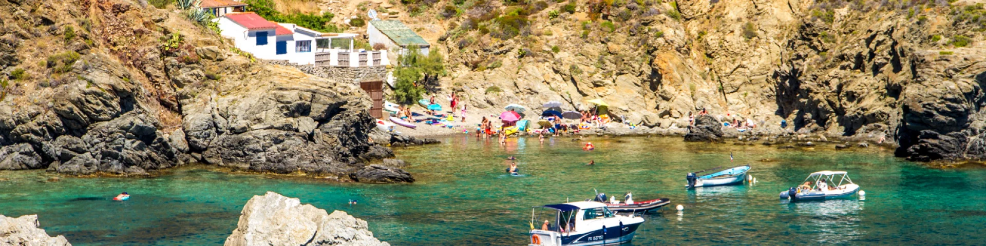 Petite crique abritée de la Côte Vermeille, bordée de rochers et de maisons blanches, où des baigneurs profitent des eaux turquoise et des bateaux mouillent près du rivage.
