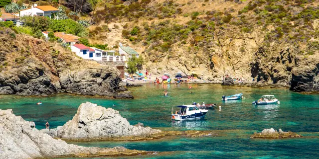 Petite crique abritée de la Côte Vermeille, bordée de rochers et de maisons blanches, où des baigneurs profitent des eaux turquoise et des bateaux mouillent près du rivage.