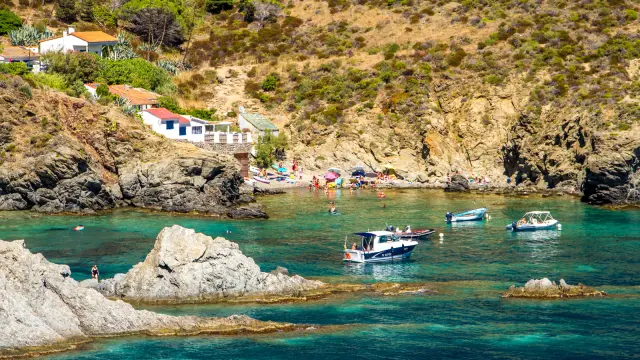 Petite crique abritée de la Côte Vermeille, bordée de rochers et de maisons blanches, où des baigneurs profitent des eaux turquoise et des bateaux mouillent près du rivage.